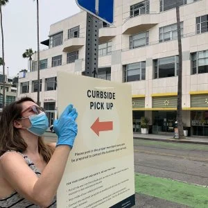 A woman in a medical mask and gloves hangs a "curbside pickup" sign.